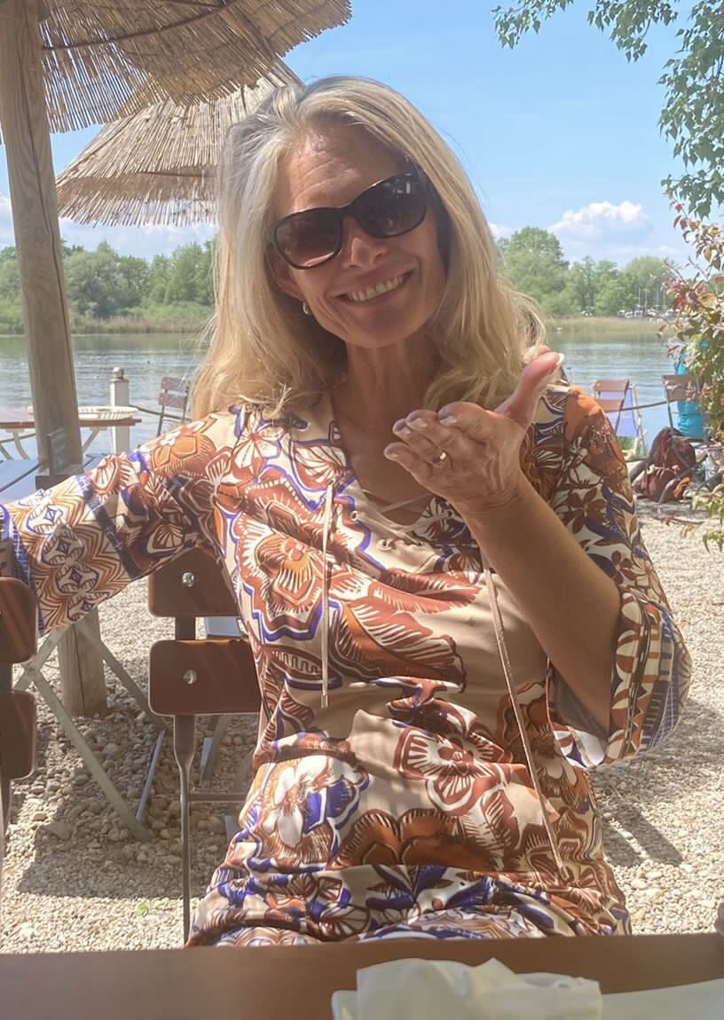 Auf dem Bild sieht man Marion Lang wie sie locker auf einem Stuhl in einer Strandbar sitzt. Man sieht im Hintergrund Gewässer einen schönen blauen Himmel und Bäume. Der Untergrund auf dem sie sich befindet ist sandig.