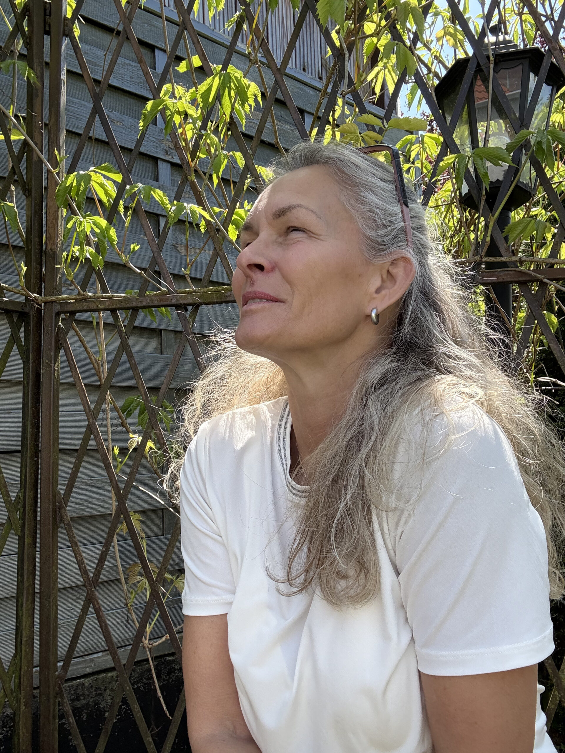 Auf dem Bild sieht man Marion Lang. Man sieht sie leicht von ihrer linken Seite. Sie trägt ihre blonden Haare offen mit einem halben Zopf nach hinten gebunden. Sie trägt einen weißen Pullover. Sie schaut von träumt in die Gegend. Im Hintergrund sieht man einen Zaun mit grünen Bewachsungen.