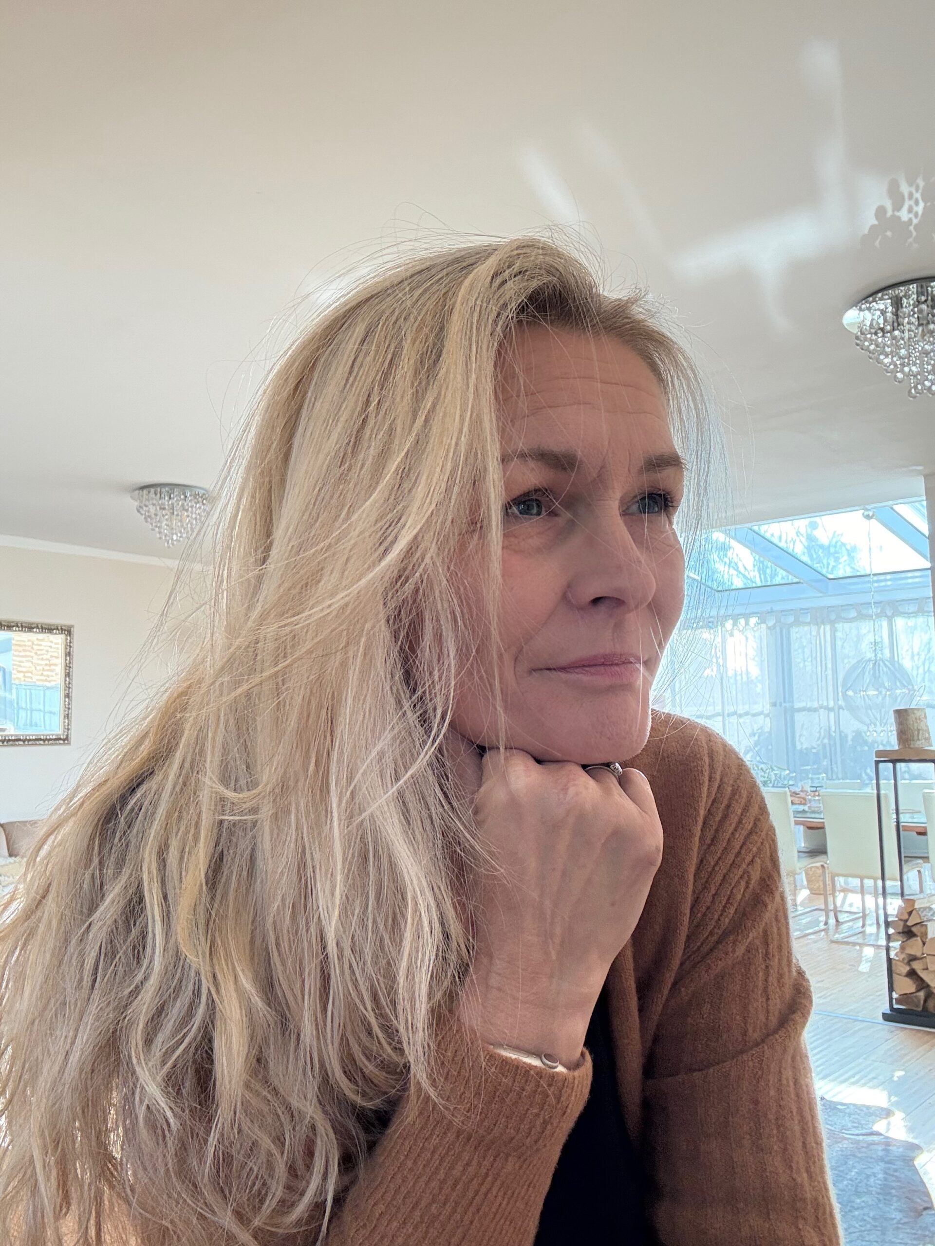 Auf dem Bild sieht man Marion Lang rechts von der Seite. Sie schaut nachdenklich und hat ihren Kopf auf ihre rechte Faust gestützt. Ihre langen blonden Haare liegen auf ihrer rechten Seite ganz locker auf. Im Hintergrund sieht man eine Wohnung wo die Sonne durch die Fenster scheint.