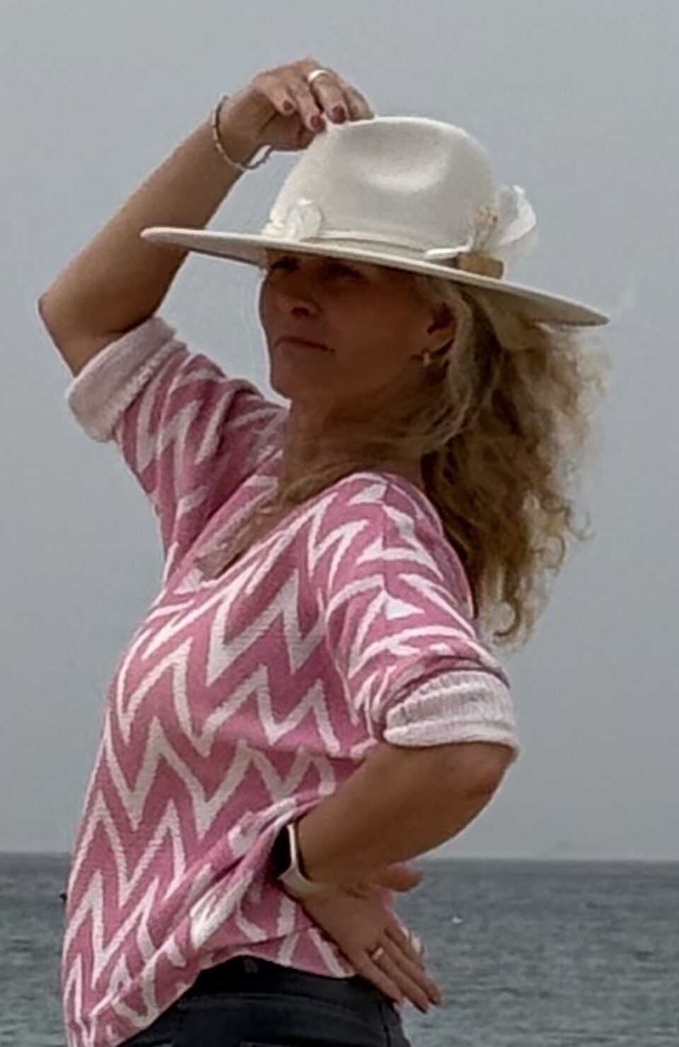 Auf dem Bild sieht man Marion Lang. Man sieht sie links von der Seite. Sie trägt keck einen weißen Hut auf dem Kopf den sie mit ihrer rechten Hand antippt. Ihre blonden Haare fallen locker nach hinten weg. Sie trägt einen rosa weiß gemusterten Pullover und im Hintergrund sieht man den Himmel.